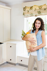 Woman holding paper bag filled with carrots celery pepper and oranges near white kitchen cabinets