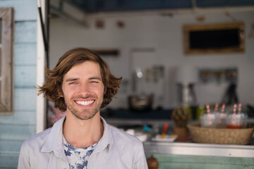 Mid adult man smiling behind pastel blue counter at cafe with blender and basket holding cups