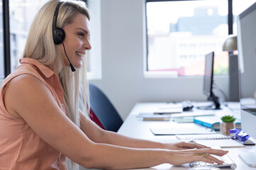 Woman sitting at desk wearing headset, smiling at computer monitor with open notebook and succulent