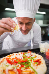Male chef wearing white toque jacket sprinkling shredded cheese onto pizza crust at kitchen counter