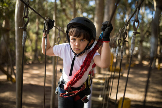 Boy navigating suspended rope course in woods wearing black helmet, gloves, red black harness - Powered by Adobe