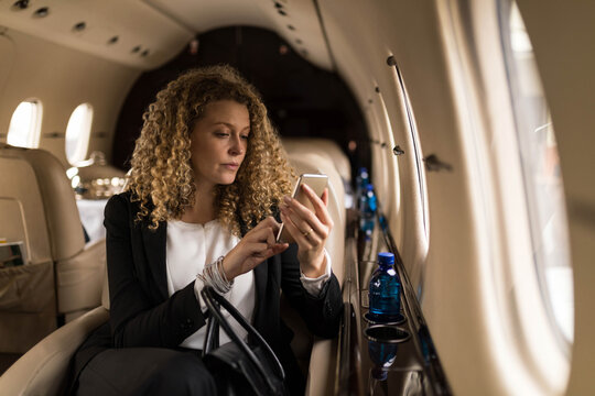 Businesswoman tapping smartphone screen in private jet cabin with blue water bottle