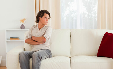 Fototapeta premium Man sitting on off-white sofa gazing right at home with red pillow, dried-flower vase, copy space
