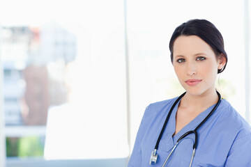 Female medical worker wearing blue scrubs holding stethoscope standing at clinic window, copy space