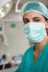 Surgeon standing in surgery room wearing scrubs with facemask and monitors glowing under lights