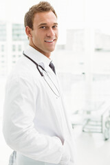 Male doctor wearing white lab coat and tie, standing near windows in clinic with stethoscope