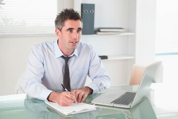 Man in dress shirt and tie writing on paperwork at glass-top desk in office with laptop