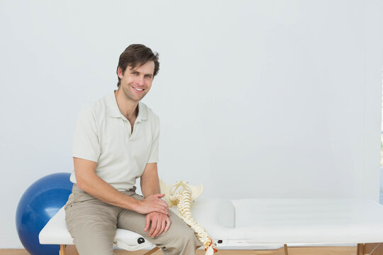 Mid adult man sitting on treatment table wearing polo shirt next to spine model, exercise ball