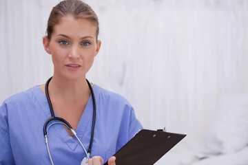 Female medical professional standing with black clipboard in exam room wearing scrubs stethoscope