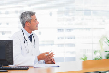 Male doctor sitting at desk by computer monitor marking clipboard wearing lab coat stethoscope