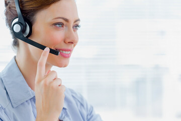 Female call center agent talking on headset, looking right at desk by window blinds, copy space