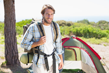 Man standing at campsite clearing holding hiking backpack with sleeping mat near pine tree