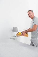 Middle-aged man standing in bedroom holding breakfast tray with juice pastry and flower, copy space