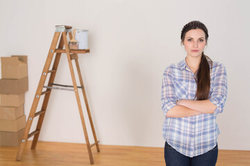 Woman standing near ladder wearing plaid shirt and jeans with paint can in room under renovation