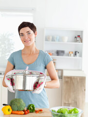 Woman standing at kitchen countertop wearing oven mitts and holding steel pot near broccoli