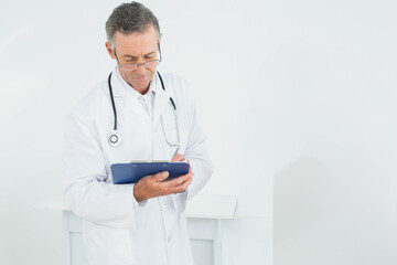 Male doctor wearing white coat with stethoscope reading notes on clipboard in clinic, copy space