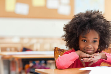 Fototapeta premium African American girl resting chin on hands at wooden desk with papers in classroom, copy space
