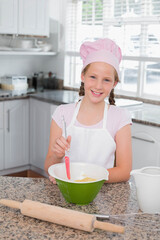 Child girl wearing pink chef hat and apron stirring batter in mixing bowl in residential kitchen