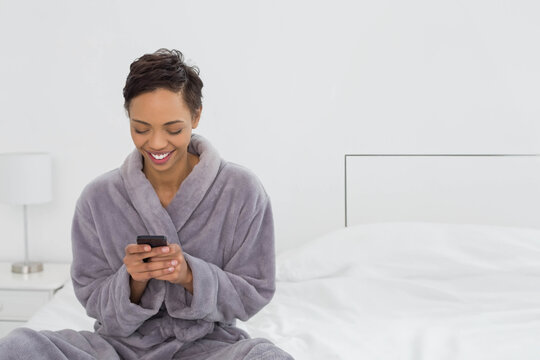 African American woman sitting on bed in bedroom wearing gray bathrobe and holding smartphone - Powered by Adobe