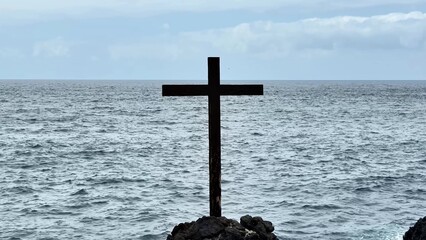 A stunning cross stands proudly on the rugged rocks by the vast ocean, symbolizing deep peace and...