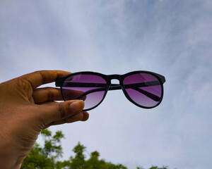 Hand Holding Black Sunglasses with Purple Lenses Against Blue Sky