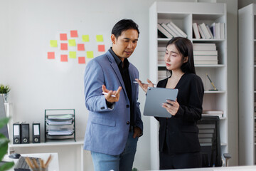 Two asian coworkers standing in the office discuss about business plan and looking at tablet.handsome asia man and beautiful asian woman as business partners working at indoor
