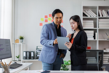 Two asian coworkers standing in the office discuss about business plan and looking at...