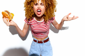 Energetic young woman with curly hair expressing frustration while holding a hamburger, wearing a...