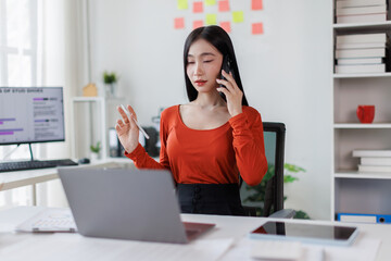 Obraz premium Asian woman working at workplace desk holds cellphone staring at laptop, using mobile phone and laptop computer Financial and Accounting concept. 