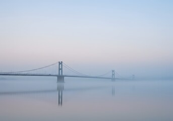 Obraz premium A suspension bridge shrouded in dense fog, reflected in the calm water below, under a pastel sky.