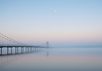 Fototapeta premium A long suspension bridge stretches across a calm, misty body of water under a pastel sky.