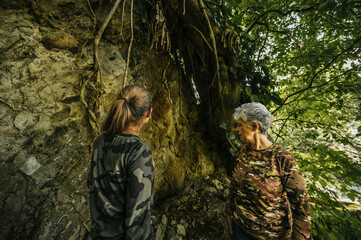Fototapeta premium Mother and daughter dressed in hiking clothes, walking next to a huge tree that stands on a giant rock in the middle of the rainforest.