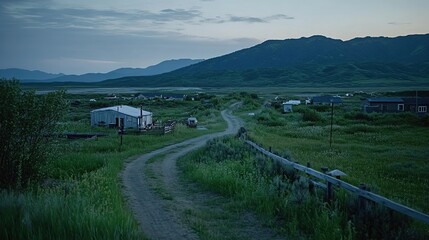 Fototapeta premium Winding dirt road through a remote village in lush green fields with distant mountains under a cloudy twilight sky