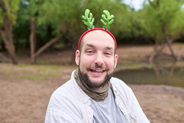 A young Hispanic man wearing a Christmas reindeer antler headband smiles at the camera outdoors, in...
