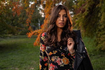 Beautiful brunette with floral dress and leather jacket posing in the park, looking at camera, very confident 
