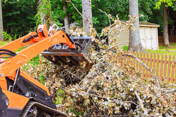 Skid steer loader removes debris from yard, clearing away fallen branches leaves in suburban area...