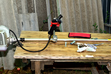 Side view of a petrol trimmer lying on a table. There is a metalworking tool next to the trimmer.
