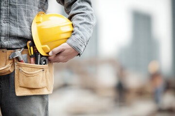 Construction worker holding his yellow safety helmet and tool belt, standing in a construction site