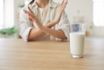 Glass of milk stands on table with woman refusing it due to lactose intolerance, a food allergy...