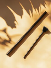 Two black, minimalist items?a long straw and a small brush?rest on a beige surface with sun-dappled shadows of leaves