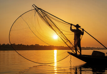 Dramatic silhouette of a lone fisherman aboard a boat, expertly throwing a large fishing net against the vibrant backdrop of a golden sunset over a calm river.