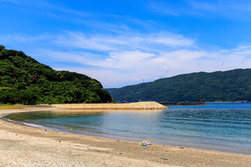 愛媛県伊方町、砂浜から望むムーンビーチ井野浦と防波堤、遠くに見える風車の夏景色

