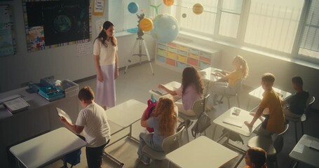 Primary School Children Preparing for Lesson, Laying Out School Supplies on Desks in Classroom. Female Teacher Greeting Group of Diverse Kids at the Beginning of School Day. Slow Motion. High Angle.