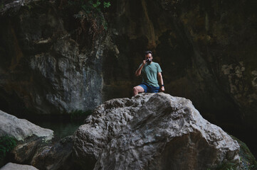 A man enjoys a warm beverage while standing on a rock, surrounded by a serene canyon with natural rock formations and lush greenery. The scene conveys peace and adventure.