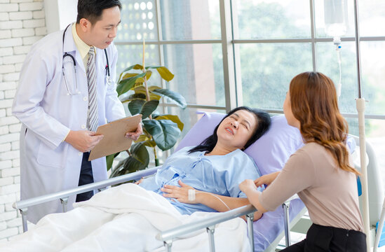 Asian man professional doctor visiting ward, consulting talking to physician, senior woman patient lying on hospital bed experiencing pain symptoms with her lovely daughter family showing care support