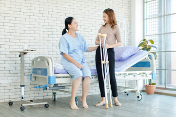 Fototapeta premium Asian young woman teaching using crutches walks beside senior female patient sitting on hospital bed medical recovery room caregiver specialist wellness healthcare treatment rehabilitation support.