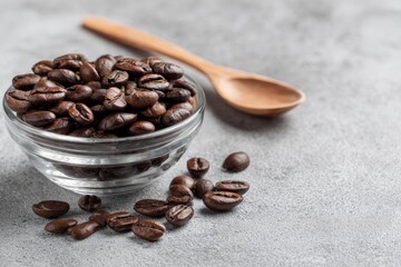Fototapeta premium Coffee beans fill a glass bowl next to a wood spoon on a gray surface Some beans are scattered nearby