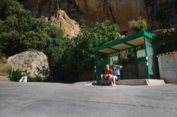 Two people sitting at a green bus stop beneath rocky cliffs, enjoying a sunny day in a picturesque rural area.