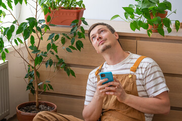 Gardener sits on the floor, holding a smartphone and looking upwards, contemplating amongst his...