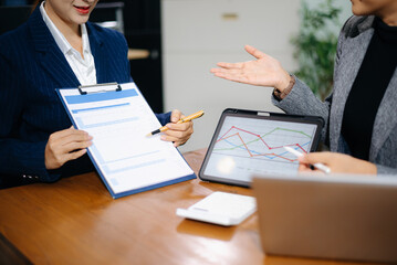 Two professional women in suits reviewing financial charts and analytics on paper and tablet in office. Business analysis, planning, and corporate strategy concept.